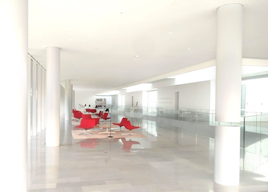 A spacious and modern interior hallway with bright lighting and minimalistic design. The area is adorned with red lounge chairs and sleek tables set upon light-colored rugs. The walls and pillars are white, enhancing the clean and open atmosphere.