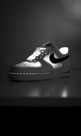 A white sneaker with a prominent black swoosh logo is placed on a reflective surface. The shoe is partially illuminated, highlighting its design features such as laces, perforations on the toe box, and a thick sole with a visible 'AIR' branding on the side. The background is a gradient of gray shades, creating a dramatic contrast.