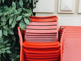 A stack of bright red plastic chairs is positioned against a beige wall with geometric patterns. To the left, green leafy plants add a natural contrast to the vibrant red of the chairs. The chairs are stacked neatly, providing a sense of order.