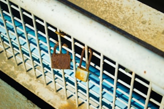Two padlocks, one rusted and one slightly newer, hang on a metal grate with a blue background visible through the bars.