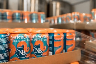 A close-up view of several cans of beer from Narrows Brewing Co., arranged in a cardboard tray. The cans feature a blue background with orange octopus tentacle graphics wrapping around the top. The branding is prominently displayed on the cans.