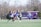 A group of girls are playing lacrosse on a grassy field. They are wearing sports uniforms and protective eyewear. The scene captures them actively running and holding lacrosse sticks, illustrating a dynamic moment of the game. In the background, there are trees and a purple building, suggesting an outdoor sports facility.