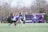 A group of girls are playing lacrosse on a grassy field. They are wearing sports uniforms and protective eyewear. The scene captures them actively running and holding lacrosse sticks, illustrating a dynamic moment of the game. In the background, there are trees and a purple building, suggesting an outdoor sports facility.
