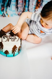 A baby in a striped shirt with a fox illustration is sitting on a white surface, reaching out towards a cake. The cake is decorated with icing to resemble a snowy landscape with brown trees and has an ombre blue base.