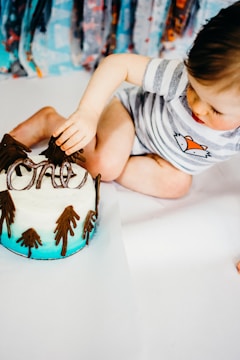 A baby in a striped shirt with a fox illustration is sitting on a white surface, reaching out towards a cake. The cake is decorated with icing to resemble a snowy landscape with brown trees and has an ombre blue base.