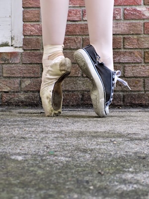 A pair of feet is shown against a brick wall. One foot is wearing a well-worn ballet pointe shoe, and the other foot is wearing a black high-top sneaker with a white sole. The contrasting footwear highlights a blend of elegance and casual style.