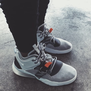 A pair of stylish, modern sneakers featuring gray and black accents with orange tags. The shoes have a sleek design, showcased on a textured concrete floor.