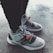 A pair of stylish, modern sneakers featuring gray and black accents with orange tags. The shoes have a sleek design, showcased on a textured concrete floor.
