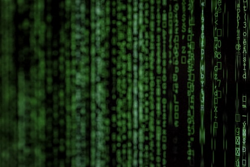 Columns of green digital code resembling binary or programming language symbols cascade down a dark background, reminiscent of a computer screen interface.