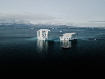 A large iceberg with a natural arch towers over a calm sea. A wooden sailing ship is positioned near the iceberg, highlighting its massive size. The background features distant snowy mountains and a cloudy sky, creating a serene and vast arctic landscape.