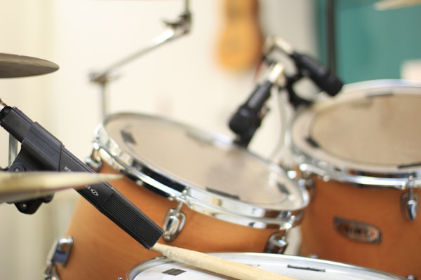A close-up view of a drum set with a focus on a wooden drumstick placed near the drumheads. Several microphones are positioned around the drums for sound capture. The drums have a natural wooden finish with chrome hardware, and the background is slightly blurred.