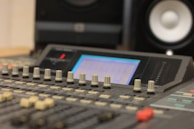A close-up view of a sound mixing console with numerous knobs and sliders. The display screen is softly illuminated, suggesting active use, and a pair of blurred speakers can be seen in the background, emphasizing a studio environment.