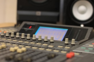 A close-up view of a sound mixing console with numerous knobs and sliders. The display screen is softly illuminated, suggesting active use, and a pair of blurred speakers can be seen in the background, emphasizing a studio environment.