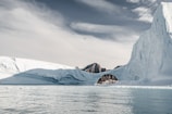 A vast icy landscape featuring a large iceberg with an arch formation, water in the foreground, and rugged, snow-covered mountains in the distance under a cloudy sky.