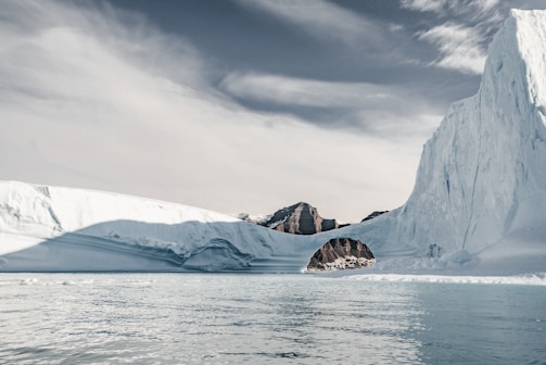 A vast icy landscape featuring a large iceberg with an arch formation, water in the foreground, and rugged, snow-covered mountains in the distance under a cloudy sky.