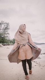 A person in a light brown hijab and coat stands on a sandy beach with a subtle smile, holding the coat open. The ocean waves are visible in the background, and trees line the coast.