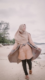 A person in a light brown hijab and coat stands on a sandy beach with a subtle smile, holding the coat open. The ocean waves are visible in the background, and trees line the coast.