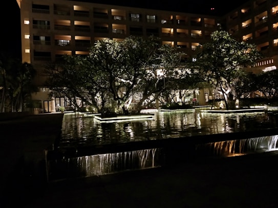 A nighttime scene with a well-lit multi-story building behind a water feature. Trees with illuminated branches are seen in the foreground, casting reflections on the water, with small waterfalls adding to the ambiance. Warm lights from the building windows provide a comforting glow.