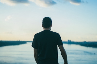A person stands overlooking a serene body of water with a subtle sky above, suggesting contemplation or peaceful solitude. The individual is viewed from behind, wearing a dark shirt, and the landscape stretches beyond with muted colors.