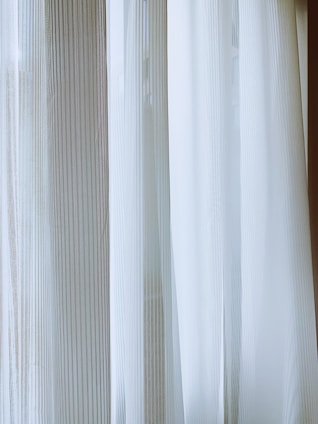 Light-colored curtains with vertical stripes hang from a window, allowing diffused natural light to pass through. The fabric has a sheer, delicate quality, creating a soft and serene ambiance.