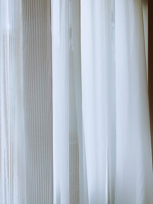 Light-colored curtains with vertical stripes hang from a window, allowing diffused natural light to pass through. The fabric has a sheer, delicate quality, creating a soft and serene ambiance.