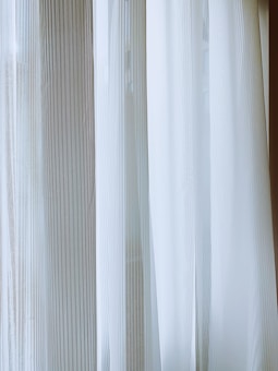 Light-colored curtains with vertical stripes hang from a window, allowing diffused natural light to pass through. The fabric has a sheer, delicate quality, creating a soft and serene ambiance.