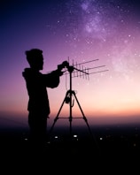 A silhouetted person adjusts an antenna on a tripod against a backdrop of a starry sky and a gradient from purple to orange at the horizon.