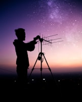 A silhouetted person adjusts an antenna on a tripod against a backdrop of a starry sky and a gradient from purple to orange at the horizon.