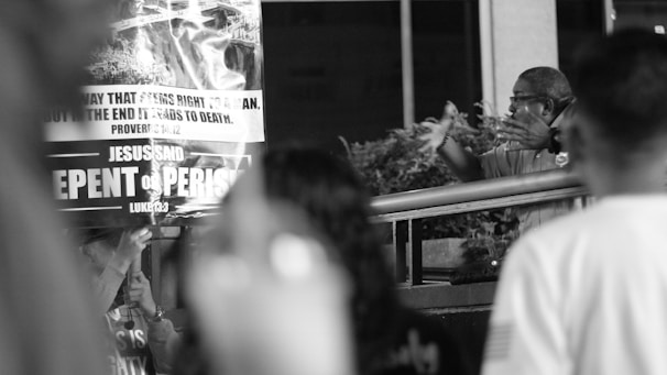 A man gestures energetically in a public setting, next to a large sign with religious messages, such as 'REPENT OR PERISH' and Bible verses.