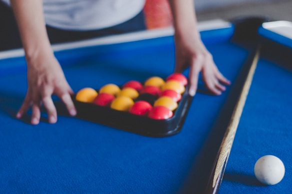 A person arranging a set of pool balls in a triangle rack on a blue pool table. The balls are brightly colored with red and yellow hues. A cue stick is placed on the right side of the table and a white cue ball rests near the edge.