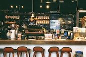 A cozy café with a warm ambiance, featuring a counter with various coffee equipment and products on display. A barista is preparing drinks behind the counter, with shelves of coffee bags and mugs in the background. The lighting is dim and atmospheric with hanging lights above.