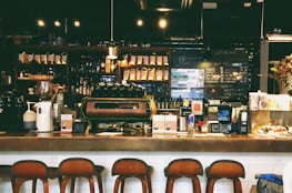 A cozy café with a warm ambiance, featuring a counter with various coffee equipment and products on display. A barista is preparing drinks behind the counter, with shelves of coffee bags and mugs in the background. The lighting is dim and atmospheric with hanging lights above.