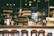 A cozy café with a warm ambiance, featuring a counter with various coffee equipment and products on display. A barista is preparing drinks behind the counter, with shelves of coffee bags and mugs in the background. The lighting is dim and atmospheric with hanging lights above.