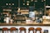 A cozy café with a warm ambiance, featuring a counter with various coffee equipment and products on display. A barista is preparing drinks behind the counter, with shelves of coffee bags and mugs in the background. The lighting is dim and atmospheric with hanging lights above.