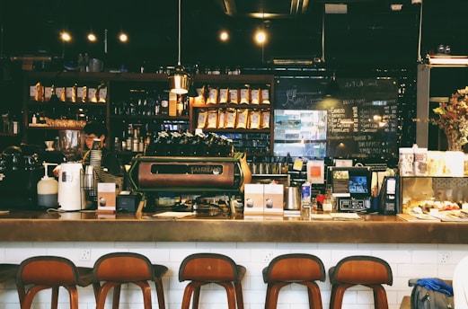 A cozy café with a warm ambiance, featuring a counter with various coffee equipment and products on display. A barista is preparing drinks behind the counter, with shelves of coffee bags and mugs in the background. The lighting is dim and atmospheric with hanging lights above.