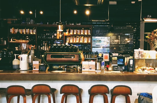 A cozy caf&eacute; with a warm ambiance, featuring a counter with various coffee equipment and products on display. A barista is preparing drinks behind the counter, with shelves of coffee bags and mugs in the background. The lighting is dim and atmospheric with hanging lights above.
