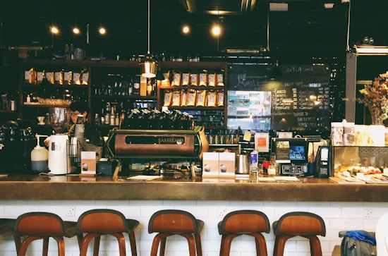 A cozy caf&eacute; with a warm ambiance, featuring a counter with various coffee equipment and products on display. A barista is preparing drinks behind the counter, with shelves of coffee bags and mugs in the background. The lighting is dim and atmospheric with hanging lights above.