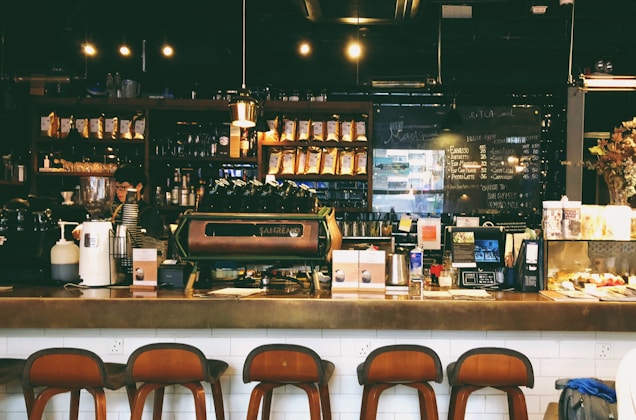 A cozy café with a warm ambiance, featuring a counter with various coffee equipment and products on display. A barista is preparing drinks behind the counter, with shelves of coffee bags and mugs in the background. The lighting is dim and atmospheric with hanging lights above.
