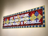 A colorful, ornate wall mural features bold text stating 'LET THE FUTURE TELL THE TRUTH. ANOTHER WORLD IS POSSIBLE.' The background consists of intricate patterns with floral and geometric designs in red, blue, yellow, and black, framed by a border with repeated circular motifs.