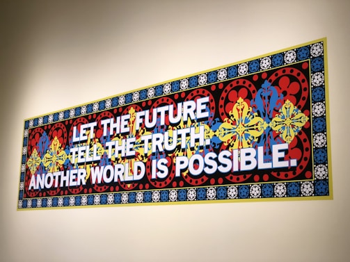 A colorful, ornate wall mural features bold text stating 'LET THE FUTURE TELL THE TRUTH. ANOTHER WORLD IS POSSIBLE.' The background consists of intricate patterns with floral and geometric designs in red, blue, yellow, and black, framed by a border with repeated circular motifs.
