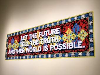 A colorful, ornate wall mural features bold text stating 'LET THE FUTURE TELL THE TRUTH. ANOTHER WORLD IS POSSIBLE.' The background consists of intricate patterns with floral and geometric designs in red, blue, yellow, and black, framed by a border with repeated circular motifs.