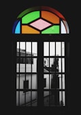 A stained glass window featuring geometric patterns with vibrant colors like green, orange, and pink. The window is set within a dark frame, casting a shadowy view of the buildings outside, which appear monochromatic and blurred.