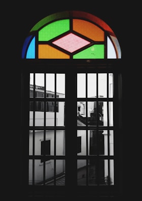 A stained glass window featuring geometric patterns with vibrant colors like green, orange, and pink. The window is set within a dark frame, casting a shadowy view of the buildings outside, which appear monochromatic and blurred.