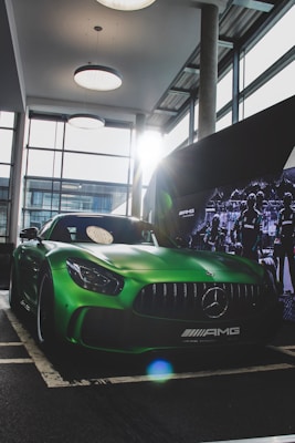 A sleek, green Mercedes-AMG sports car is parked indoors at a showroom. The car features an aggressive front grille and distinct headlights. Sunlight streams in through large windows, creating highlights and reflections on the car's surface. Surrounding the car, there are modern industrial design elements, including hanging lights and a promotional display backdrop.