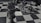 Close-up of a dark wooden chess king piece standing prominently in the foreground on a checkerboard pattern surface, with several other chess pieces blurred in the background lying on their sides.