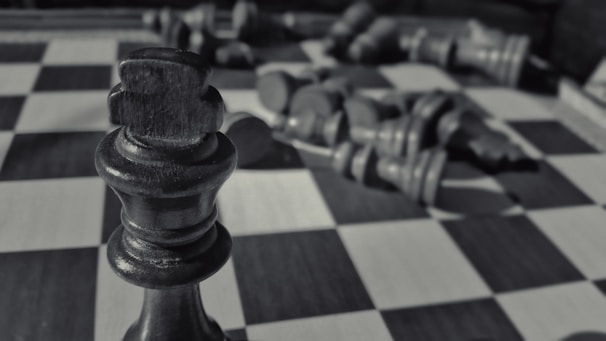 Close-up of a dark wooden chess king piece standing prominently in the foreground on a checkerboard pattern surface, with several other chess pieces blurred in the background lying on their sides.
