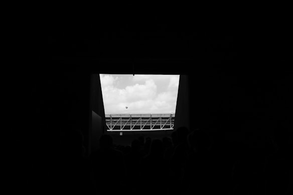 A black and white photograph depicting a group of silhouetted people standing in a dark area, looking out towards a bright opening. Beyond the opening, a soccer ball is suspended in mid-air against a backdrop of cloudy skies. A section of a modern stadium structure is visible in the distance.