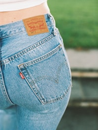 A close-up view of a person wearing a pair of blue Levi Strauss & Co. jeans, showcasing the iconic brown leather label on the waistband and a red tag on the pocket. The background features a blurred green area, possibly grass.
