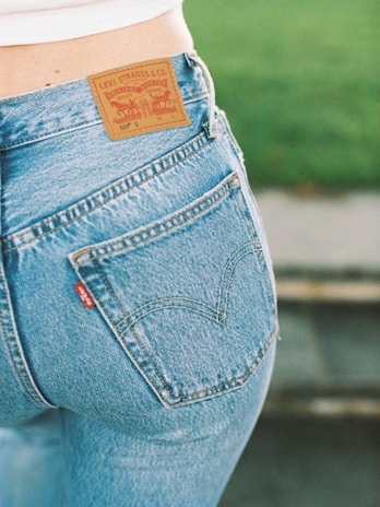 A close-up view of a person wearing a pair of blue Levi Strauss & Co. jeans, showcasing the iconic brown leather label on the waistband and a red tag on the pocket. The background features a blurred green area, possibly grass.