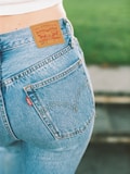 A close-up view of a person wearing a pair of blue Levi Strauss & Co. jeans, showcasing the iconic brown leather label on the waistband and a red tag on the pocket. The background features a blurred green area, possibly grass.
